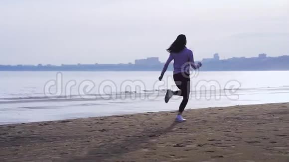 一个夏天的早晨穿着运动制服的年轻苗条的黑发女人沿着沙岸奔跑她在跑步时练习跳跃视频的预览图
