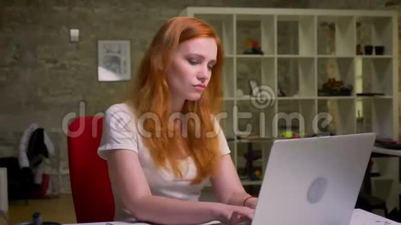 令人敬畏的红发白种人女性正坐在桌面上跳舞在她的膝上打字注意力集中视频的预览图