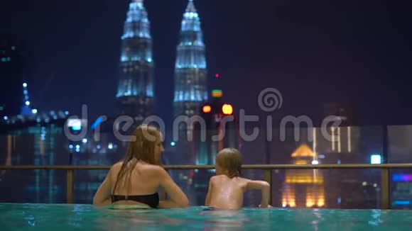 一位美丽的年轻女子和她的小儿子在屋顶的游泳池里玩得很开心可以看到摩天大楼前往亚洲视频的预览图