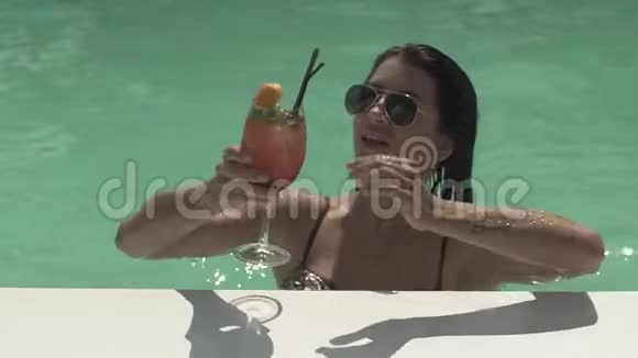戴太阳镜的布鲁内特女士正站在游泳池里喝她的鸡尾酒穿比基尼的孤独女士的休闲视频的预览图