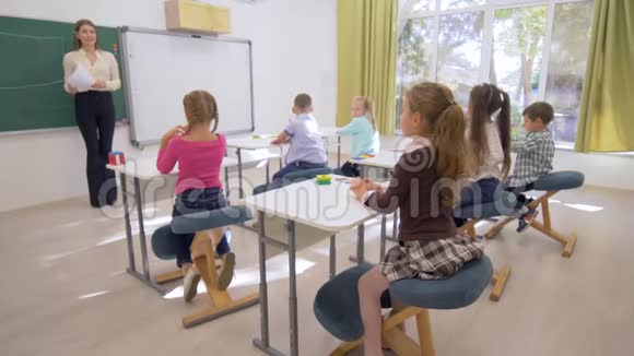 孩子们学习附近的年轻教师女士在教室的课桌上为漂亮的学童上认知课视频的预览图