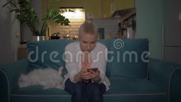 一个年轻的女人晚上和一只猫坐在沙发上用电话女孩在电话里读到了好消息视频的预览图