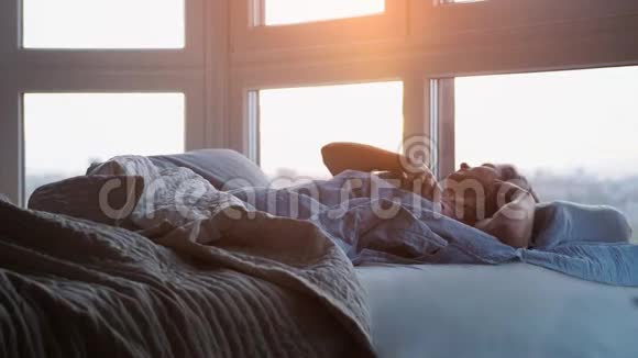 美丽浪漫的黑发女人躺在她房间的床上藏着毯子在日出时醒来慢动作视频的预览图
