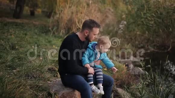 幸福的一家人和小儿子坐在秋天公园的湖岸上欣赏大自然视频的预览图