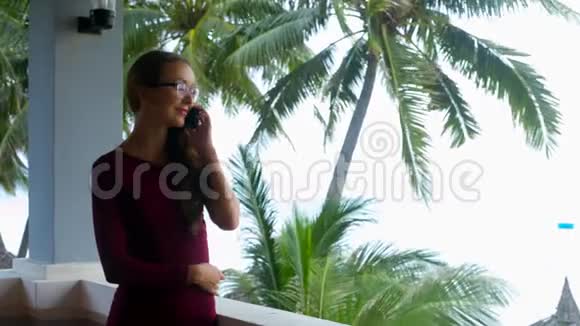 戴眼镜的女孩站在棕榈树的阳台上打电话视频的预览图