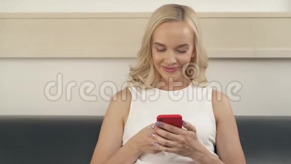 一个可爱的金发女郎坐在沙发上看着电话微笑着视频的预览图