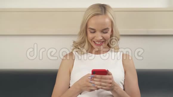 一个迷人的金发女人拿着电话坐在沙发上她看着屏幕打字她的脸视频的预览图