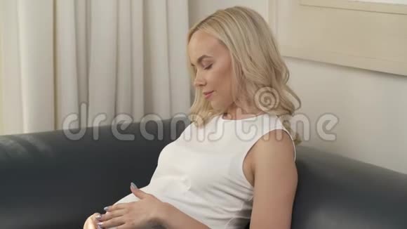 坐在沙发上的一个美丽的怀孕金发女郎的侧视图她在抚摸她的肚子看着它她停下来视频的预览图