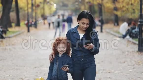 一位美丽的妈妈和她可爱的小女儿在秋天的公园里散步他们用智能手机笑着玩视频的预览图