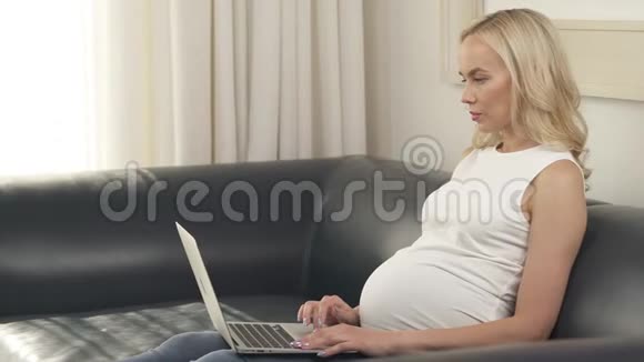 一个漂亮的怀孕金发美女坐在沙发上用笔记本电脑的侧视图她看着屏幕打字视频的预览图