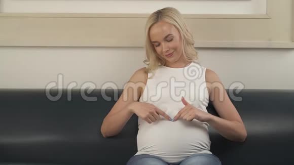坐在沙发上的一个怀孕的金发女郎的正面景色她正在用手指追踪腹部的形状她就把视频的预览图