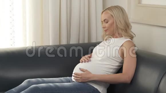一个怀孕的金发美女坐在沙发上抚摸着她的肚子她看着微笑着视频的预览图