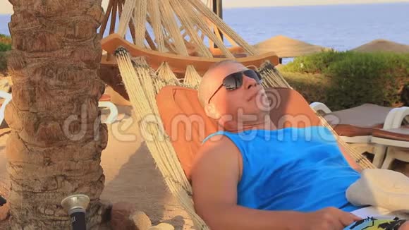 在一个夏日阳光明媚的日子里一个戴太阳镜的年轻人正躺在棕榈树荫下的海滨吊床上休息视频的预览图