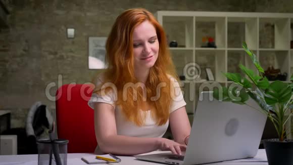 白种人生姜女孩幸福美丽的微笑坐在她的桌面上在工作期间使用笔记本电脑是个自营职业者视频的预览图