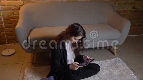 白种人女孩一头深色波浪发坐在地板上在舒适的家庭氛围中专注地看着平板电脑视频的预览图
