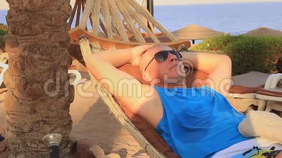 一个阳光明媚的夏日戴着太阳镜的年轻人在海滩上的吊床上休息视频的预览图