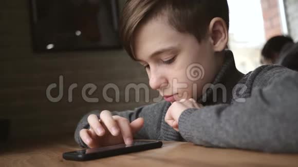 十几岁的男孩坐在窗边的咖啡馆里手里拿着一部智能手机社交网络中的交流运动会视频的预览图