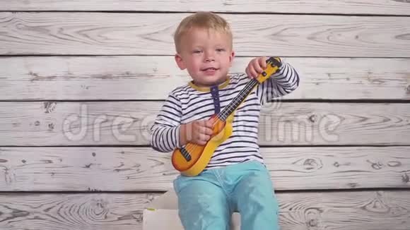 快乐的两岁男孩弹吉他或四弦琴唱歌坐在木房里视频的预览图