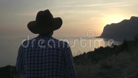 一个戴着帽子的年轻人正在海上看日落太阳落山在海角后面对夏季旅行的赞赏视频的预览图