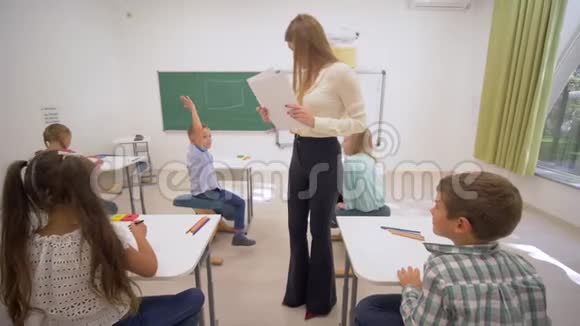 女青年教育工作者在上课期间向课桌旁的学童分发白纸进行知识检查视频的预览图