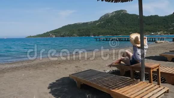 穿着衬衫戴着帽子的女孩坐在沙滩上的遮阳伞下视频的预览图