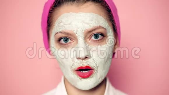 戴着化妆面具的女孩头上戴着毛巾对着粉红色的背景微笑美丽的女孩戴着化妆面具视频的预览图