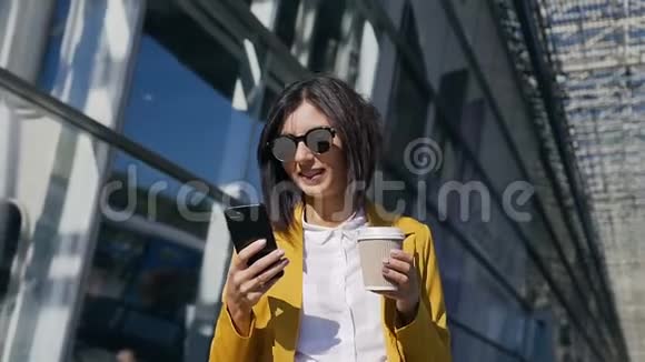 年轻的戴墨镜的职业女商人走在城市的街道上喝着咖啡用现代智能手机视频的预览图