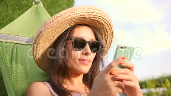 在自然日落公园的吊床上使用移动电话的太阳镜和草帽的年轻混合种族旅游女孩视频的预览图