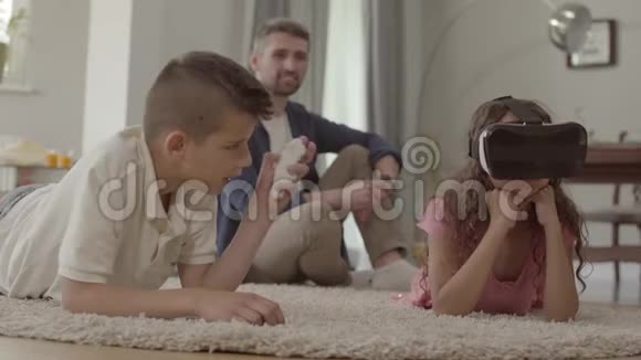 男孩和女孩用虚拟现实眼镜躺在毛茸茸的地毯上父亲坐在地毯上视频的预览图