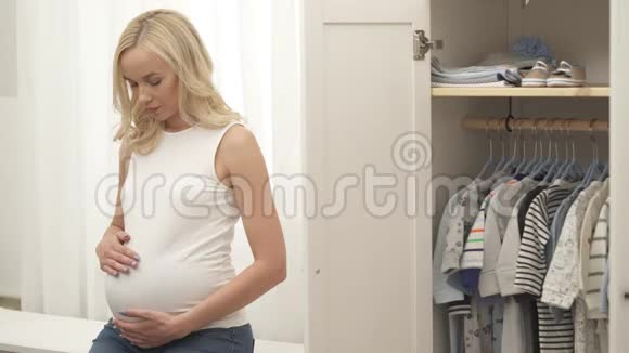 一个漂亮的怀孕金发女郎正在上下抚摸她的腹部她看着相机开心地笑着低头看着视频的预览图