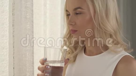 一幅站在窗边的迷人金发女郎的侧景肖像她拿着一杯水做嘴唇视频的预览图