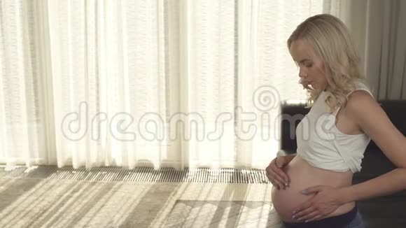 一个怀孕的金发女郎站在房间里把奶油揉进肚子里的侧视她看着肚子用视频的预览图