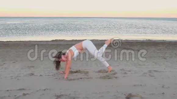 年轻女子正在做瑜伽阿多穆卡什瓦纳萨纳抬起脚在沙滩上日出视频的预览图