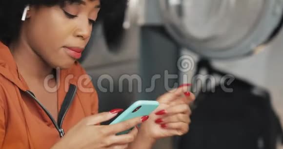 微笑着年轻的非洲裔美国妇女使用智能手机在自助洗衣店她听音乐唱歌自助服务公众视频的预览图