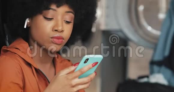 微笑着年轻的非洲裔美国妇女使用智能手机在自助洗衣店她听音乐唱歌自助服务公众视频的预览图