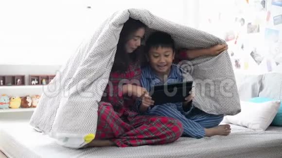 年轻女子妈妈和儿子一起在毯子下面的平板电脑上看电影视频的预览图