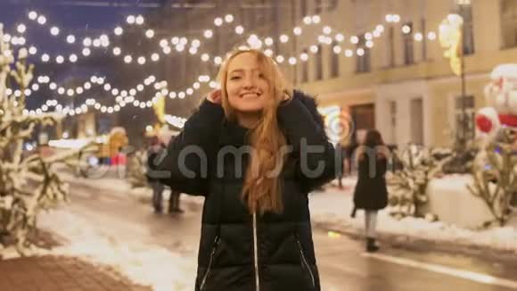 穿着冬季夹克的漂亮女孩站在冬季街道上闪烁的花环的背景上摩擦着她视频的预览图