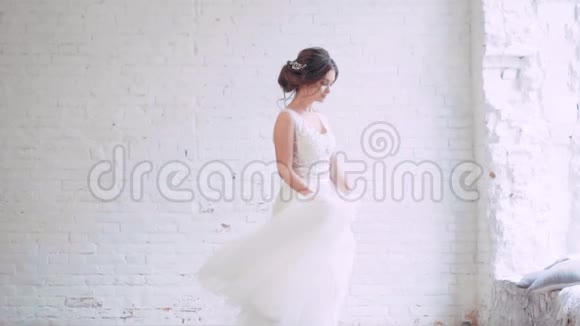 优雅的新娘穿着白色长裙的女孩在靠近柔和灰色的窗户的光线下在宽敞的房间里旋转视频的预览图