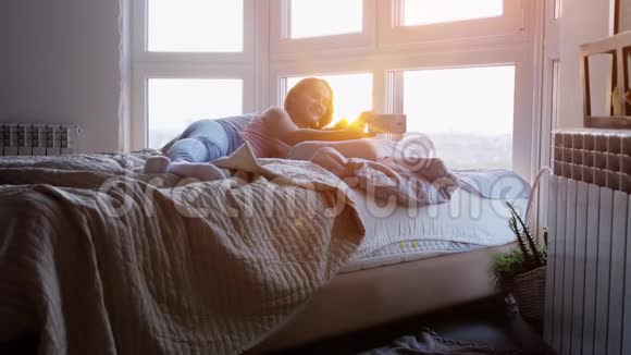 年轻快乐的女人躺在床上带着手机和缅因州的浣熊猫在日落时靠窗自拍视频的预览图