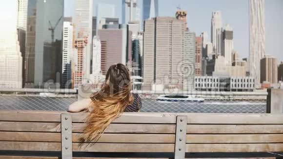 后景的快乐女孩与长发享受惊人的曼哈顿天际线景观坐在长凳上手臂张开视频的预览图