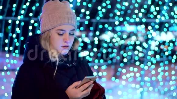 戴帽子的年轻漂亮女孩在冬天在街上使用智能手机她站在蓝色的灯光下视频的预览图