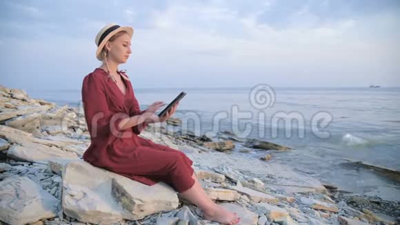 晚上一个穿着夏红色连衣裙和草帽的漂亮姑娘坐在海边的一块石头上看着视频的预览图