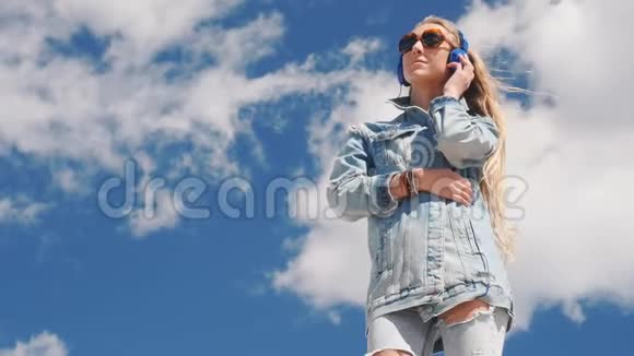 戴墨镜和牛仔裤的年轻女子在户外戴耳机听音乐视频的预览图