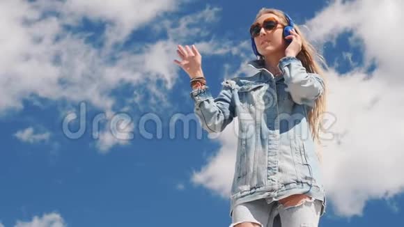 戴墨镜和牛仔裤的年轻女子在户外戴耳机听音乐视频的预览图
