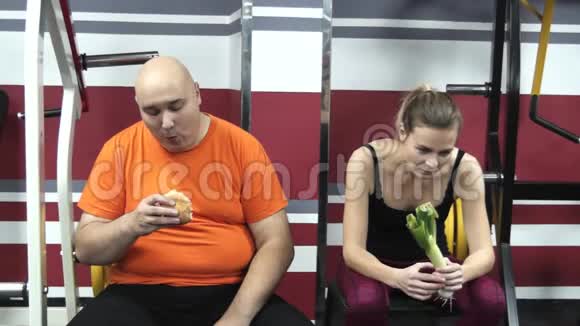 胖滑稽的男人正在健身房里吃谦卑的东西靠近他的年轻漂亮的韭菜女孩视频的预览图