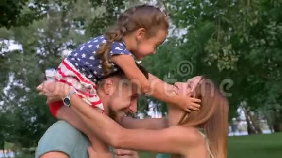 年轻的一家人夏天在公园散步女孩坐在爸爸肩膀上拥抱妈妈视频的预览图