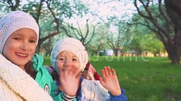两个快乐的微笑女孩坐在公园里挥动双手对着镜头在开花花园的背景下的孩子视频的预览图