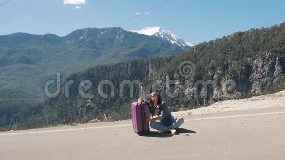 美丽的年轻女士疲惫地坐在乡村道路上在阳光明媚的日子里搭便车视频的预览图