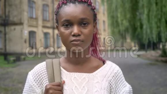 一个严肃美丽的非洲学生带着粉红色的恐惧看着相机站在大学附近的公园里视频的预览图