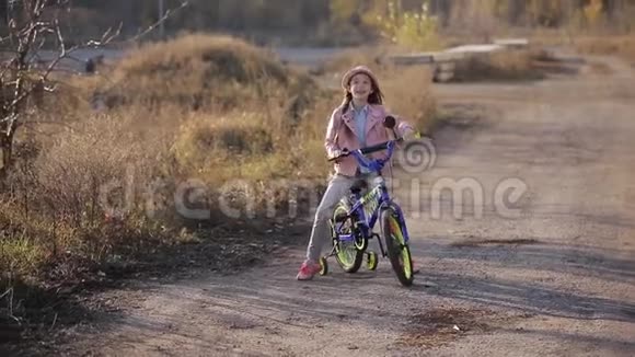 戴帽子的漂亮小女孩在夕阳的温暖光线下骑自行车在路上视频的预览图